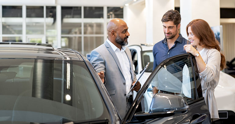 A couple of consumers are looking at a car and speaking with a representative at a dealership