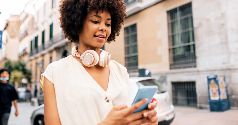 Image of a woman standing near a car with modern headphones hooked around her neck looking at her smart phone.