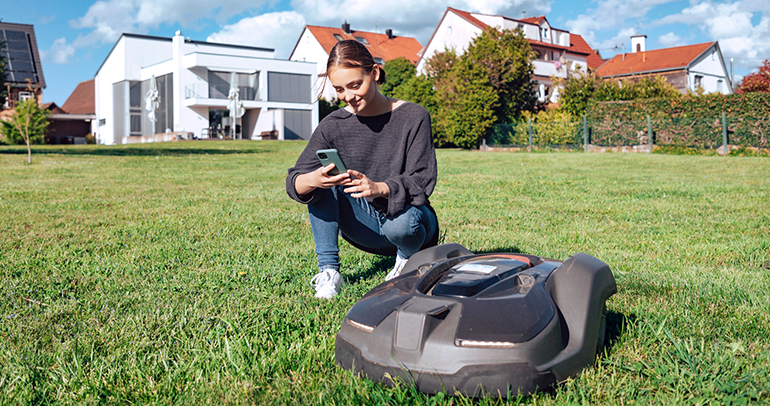 A woman kneeling in the grass is using her smartphone to control her electric-powered lawn mower that is next to her