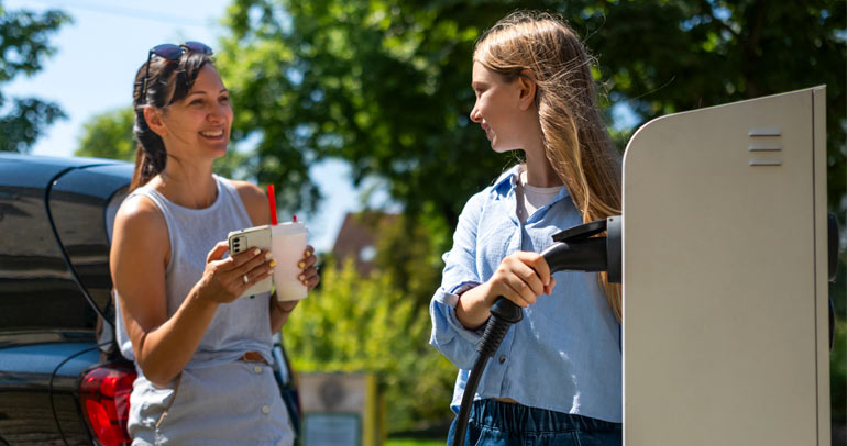 A battery electric vehicle (BEV) owner speaks with a friend as they stand outside of the BEV at a public charging station