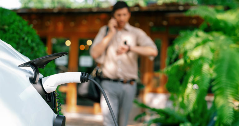 A young man is on his phone, checking his wrist watch while walking toward his his electric vehicle (EV) that is charging at a public charging station