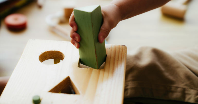 A child's hand trying to unsuccessfully fit a green triangular wooden block into a square hole