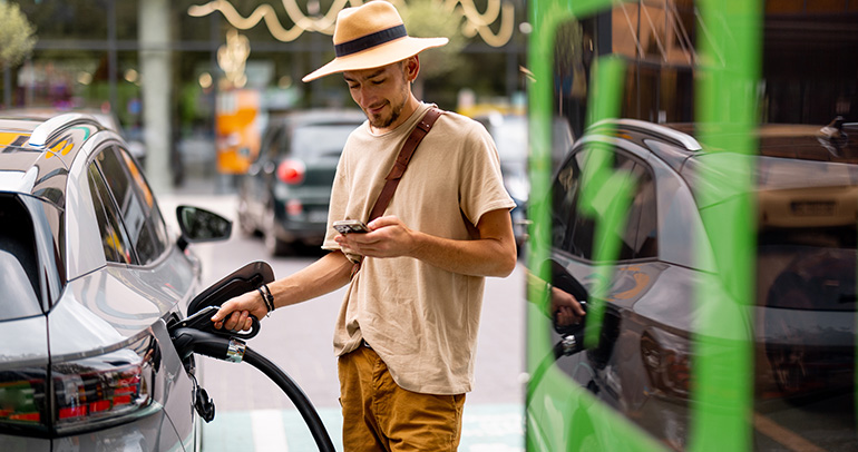 A man looking at his phone while charging his battery electric vehicle at a public fast charger