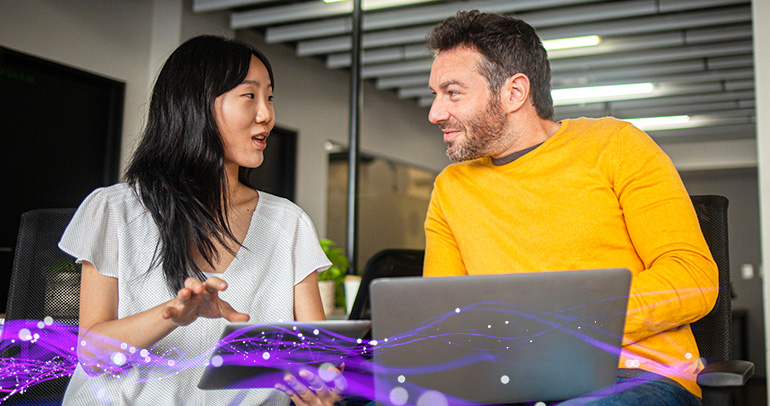 A male and female coworker are sitting side by side at a desk looking at one another and discussing what is on the laptop in front of the man and the tablet that the woman is holding. There are purple lines with light purple circles coming out of the tablet and spreading in front of the two devices, alluding to AI data.