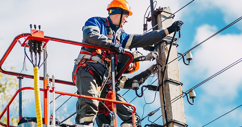 Two professional electric utility line workers in hard hats are repairing power lines from the cradle of a bucket truck