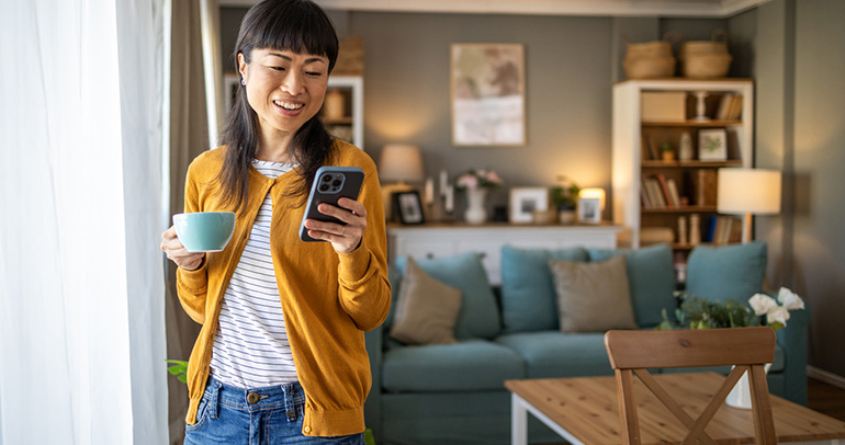 A woman at home with a cup of coffee in one hand and looking at her phone in the other hand