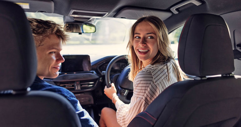 A woman in the UK sits in the right-handed driver's seat with her hands on the steering wheel and looks over her left shoulder to the back of the car while a man in the passenger seat looks over his right shoulder towards the backseat