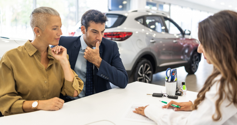 A couple sitting down at a desk in a dealership talking over the price of an electric vehicle (EV) with a car sales rep